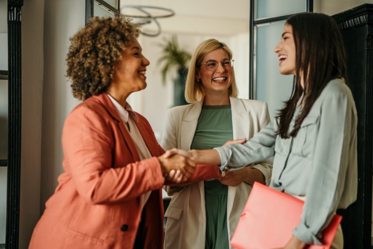 3 women smiling. 2 are shaking hands.